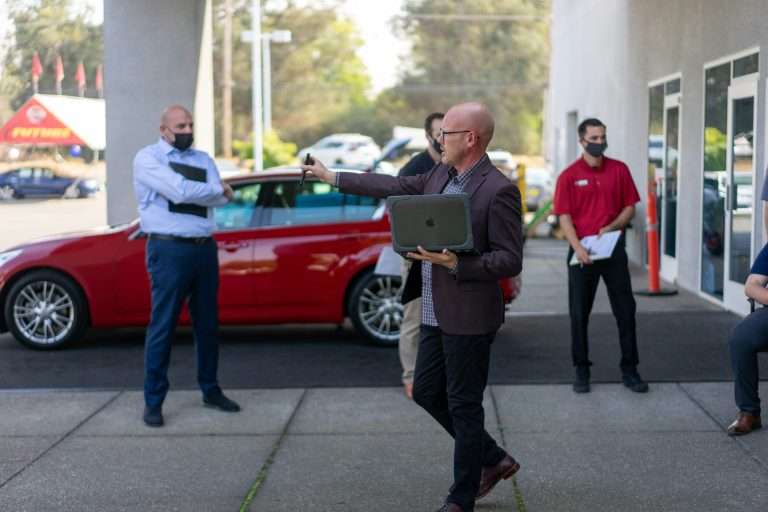 A man presenting to a group of people on the forecourt of a car dealership holding a laptop. For the RTS Academy "Foundations for Effective Management" course.