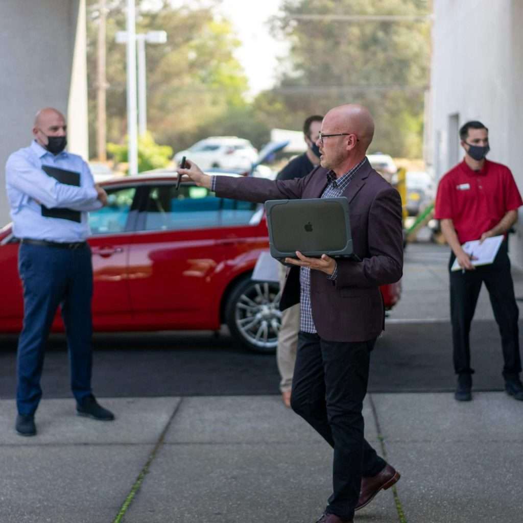 A man presenting to a group of people on the forecourt of a car dealership holding a laptop. For the RTS Academy "Foundations for Effective Management" course.