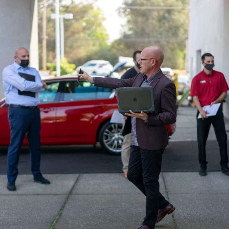 A man presenting to a group of people on the forecourt of a car dealership holding a laptop. For the RTS Academy "Foundations for Effective Management" course.