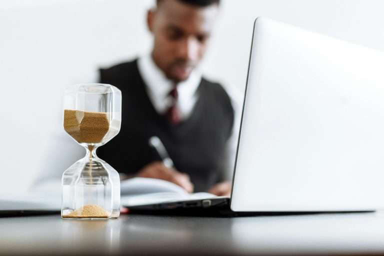 An hour glass on a desk next to a man at a computer for the RTS Academy "Effective Time Management for Leaders" and "Utilise Time for Stronger Leadership" course.
