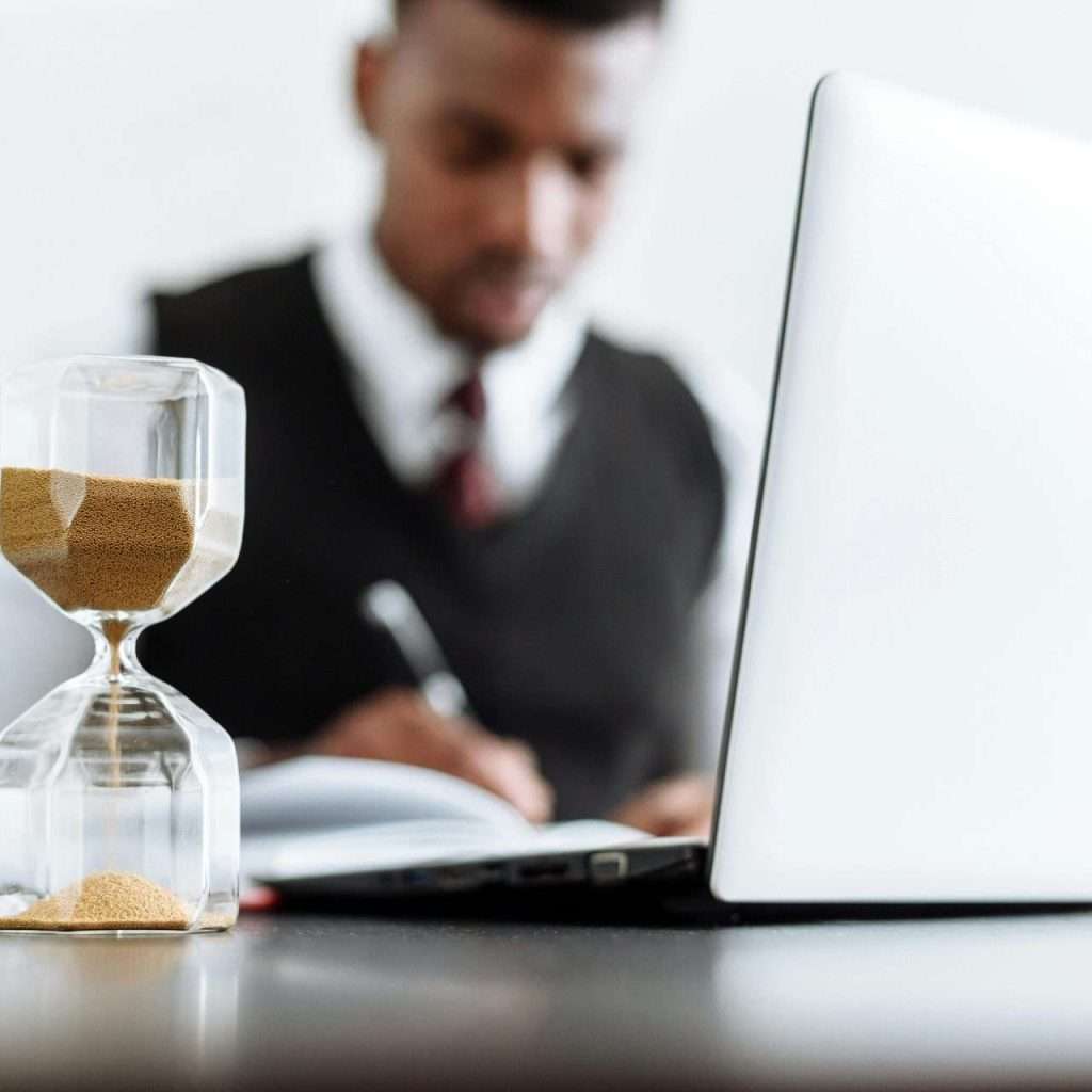 An hour glass on a desk next to a man at a computer for the RTS Academy "Effective Time Management for Leaders" and "Utilise Time for Stronger Leadership" course.