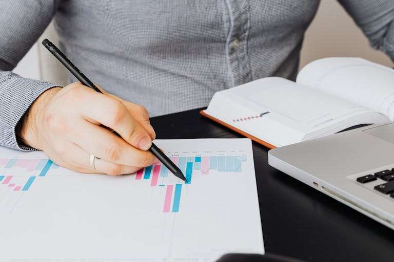 A business analyst examines a graph on paper with a laptop and notebook indoors.