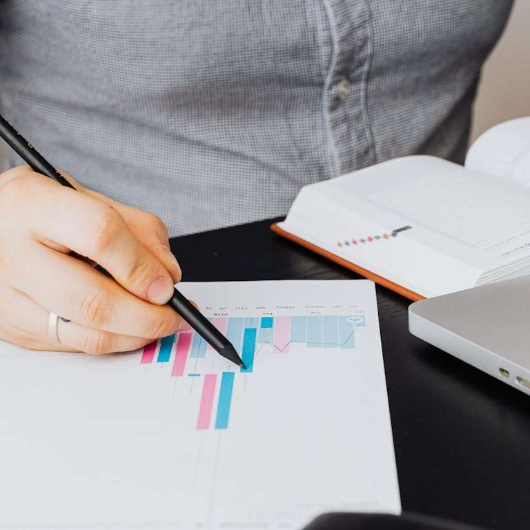 A business analyst examines a graph on paper with a laptop and notebook indoors.