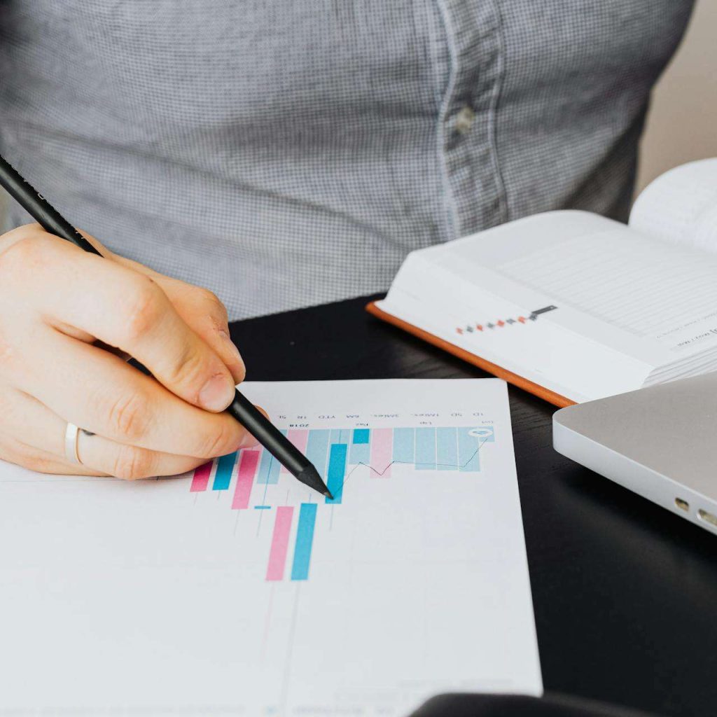A business analyst examines a graph on paper with a laptop and notebook indoors.