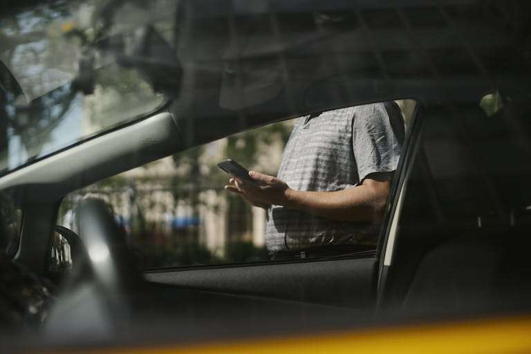 A man using a smartphone inside a car, visible through the window in an urban setting. For RTS Academy Used Vehicle Sales course.