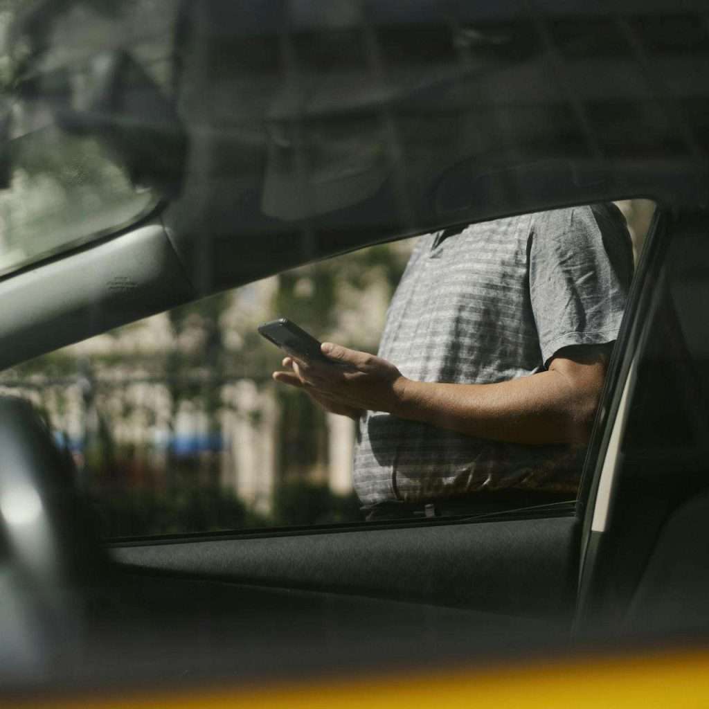 A man using a smartphone inside a car, visible through the window in an urban setting. For RTS Academy Used Vehicle Sales course.